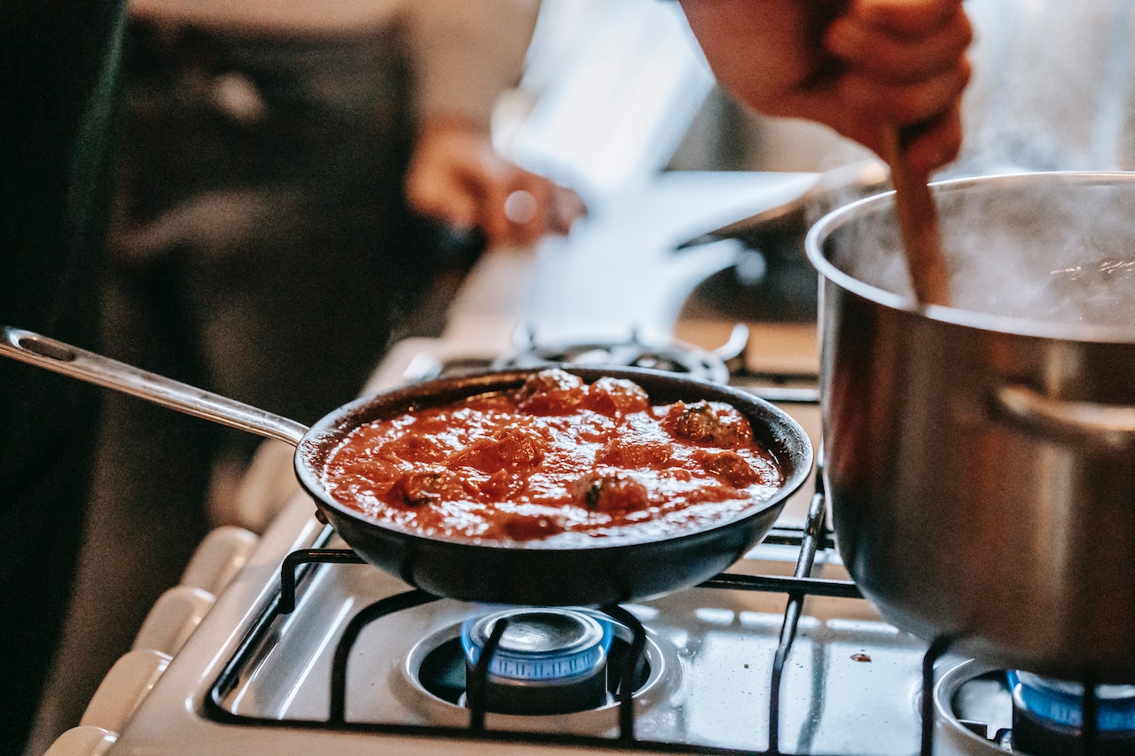 People cooking meatball in tomato sauce and pasta over gas stove