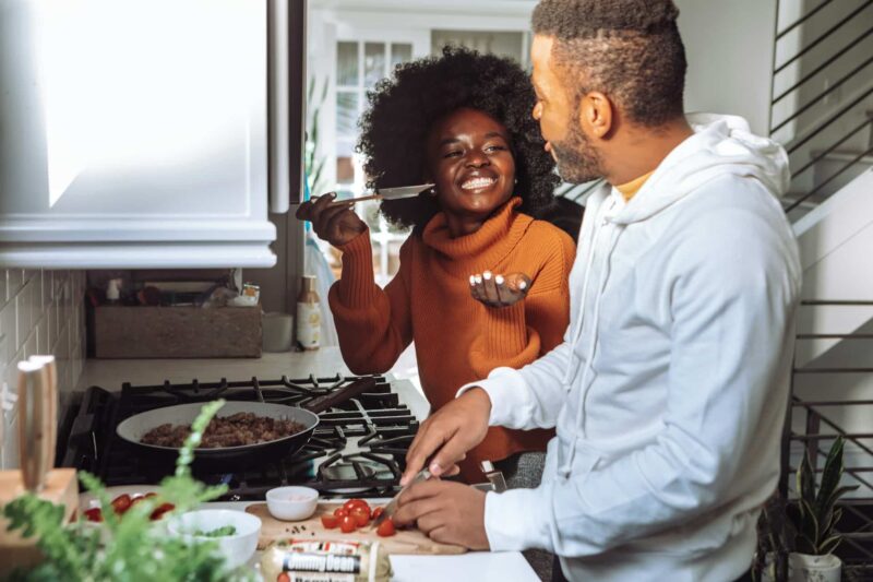 Two people smile as they cook together in a small kitchen. Improving your home's EPC rating cuts the cost of regular household bills.