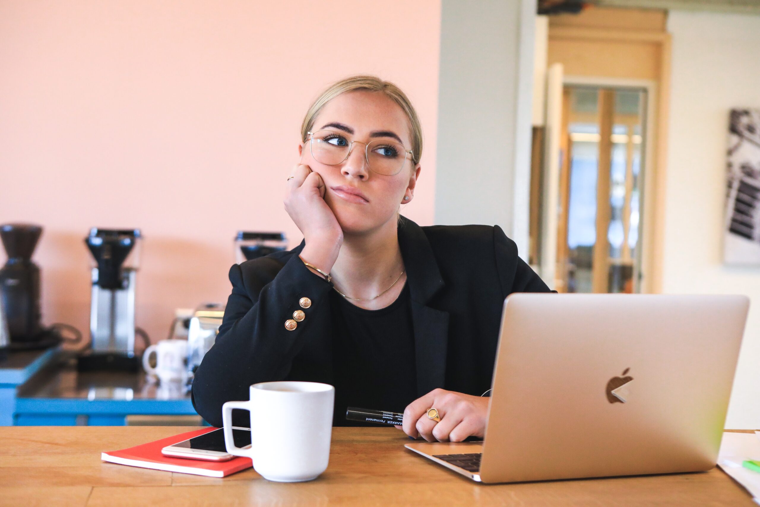 Woman in suit jacket resting her head on her hand looking frustrated with her laptop opened and a cup of beverage in front of her