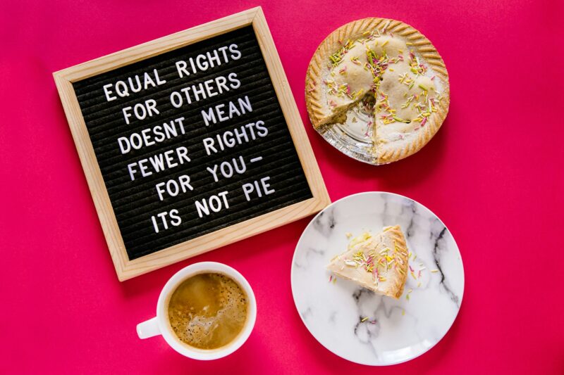 A black message board with white lettering spelling out 'Equal rights for others doesn't mean fewer rights for you - It's not pie. The message board is lying on a red table cloth with a pie with a slice cut out and a cup of tea sharing the table.